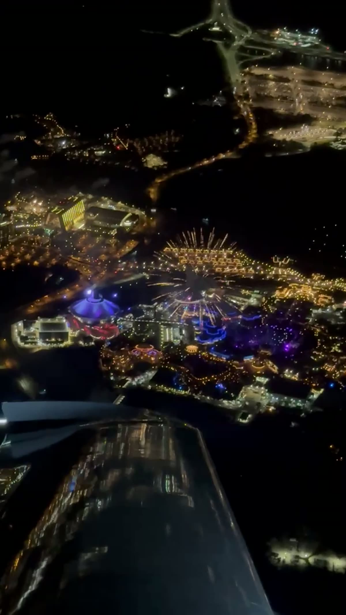 Fireworks bursting over illuminated theme parks, seen from the aircraft at night